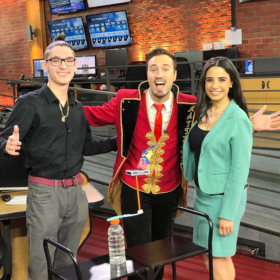 Aaron Matthews, dressed in a vibrant red and gold magician’s outfit, shares a fun moment with CP24 Breakfast hosts in Toronto. Smiling and full of energy, he showcases a magic trick with a floating object, engaging the morning show audience.