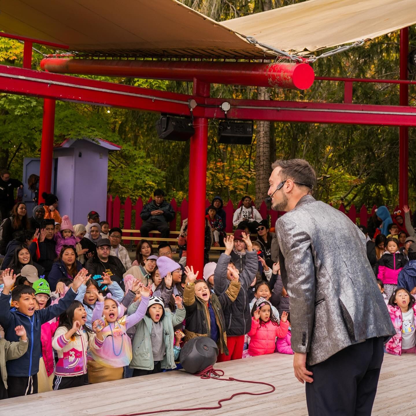 Aaron Matthews, wearing a shiny silver blazer, performs an engaging magic show for an excited crowd of children at an outdoor theater. The kids eagerly reach out with joy as Aaron interacts with them, creating a magical and fun-filled atmosphere.