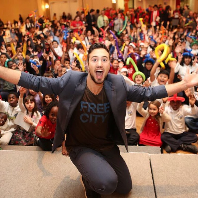 Aaron Matthews kneels on stage with arms wide open, enthusiastically engaging with a cheering crowd of children and families. Dressed in a stylish blazer and a “Free City” t-shirt, he brings excitement and joy to the audience at a live event.