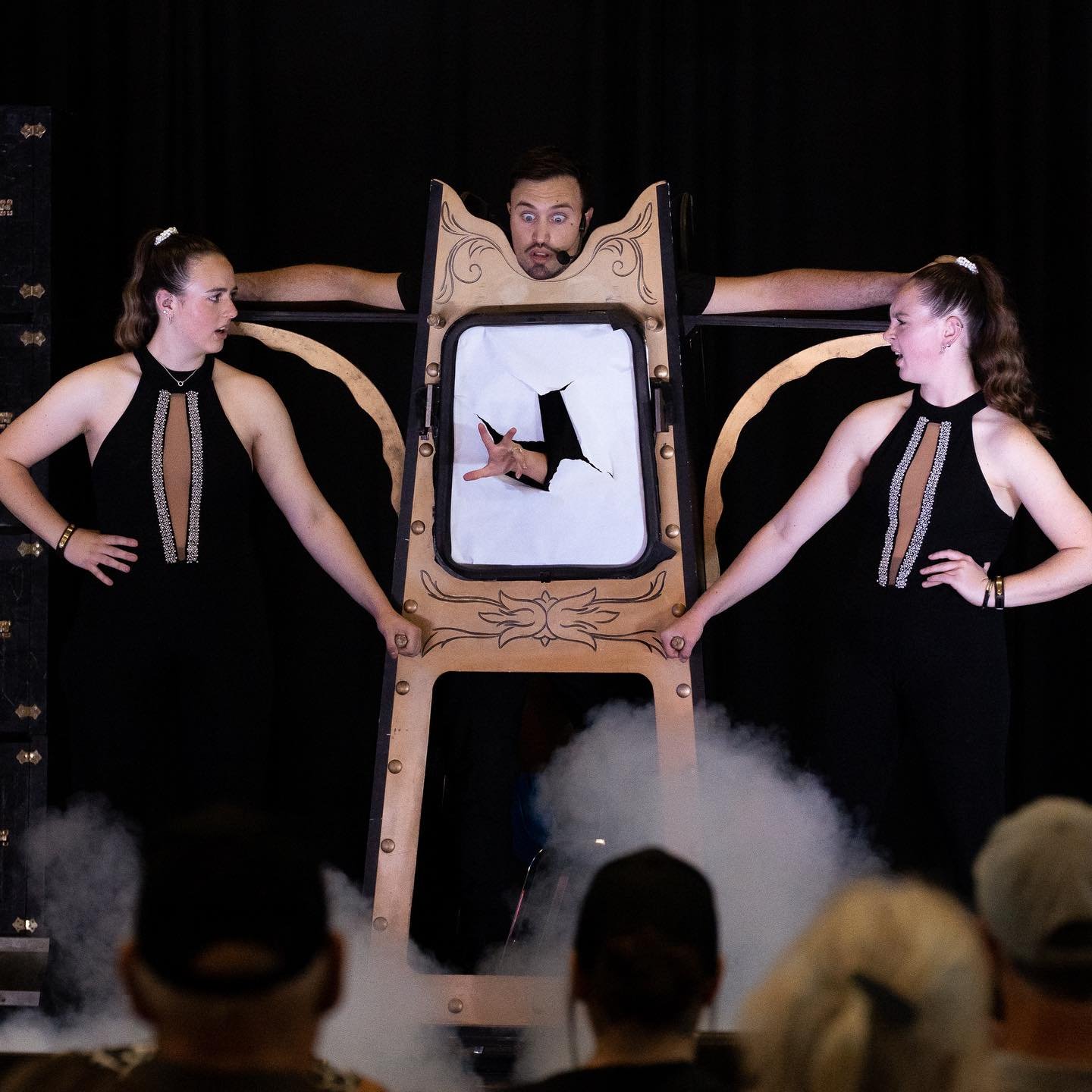 Aaron Matthews performs a thrilling stage illusion, appearing trapped inside a decorative frame with a hole in the center. Two assistants in black outfits hold the frame as smoke fills the stage, adding to the dramatic effect of the magic act.