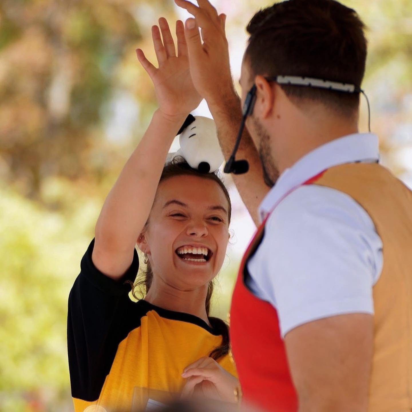 Aaron Matthews high-fives a smiling audience member during a live magic performance. The young woman, wearing a yellow and black outfit with a Snoopy plush on her head, laughs with excitement as they interact on stage.