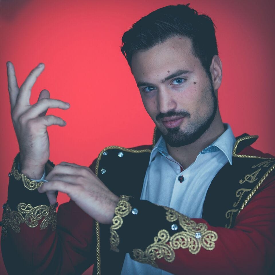 Aaron Matthews poses dramatically in a red and gold magician’s jacket against a bold red background. With intense eyes and a confident expression, he gestures with his hands, creating an aura of mystery and magic.