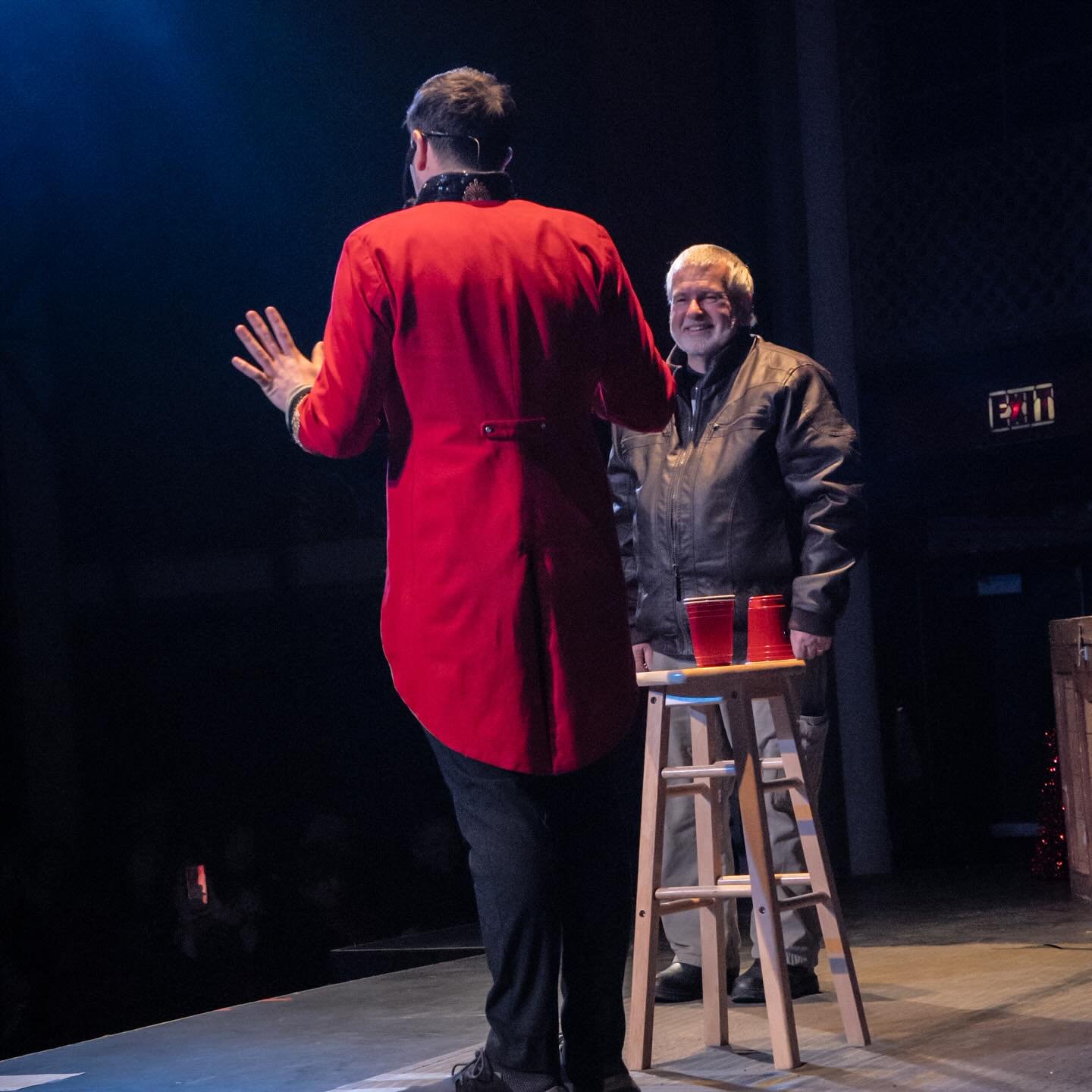 Aaron Matthews, dressed in a red magician’s coat, performs a thrilling magic trick on stage with an audience member. The guest, smiling, stands next to a stool with red cups, as the crowd eagerly watches the performance.