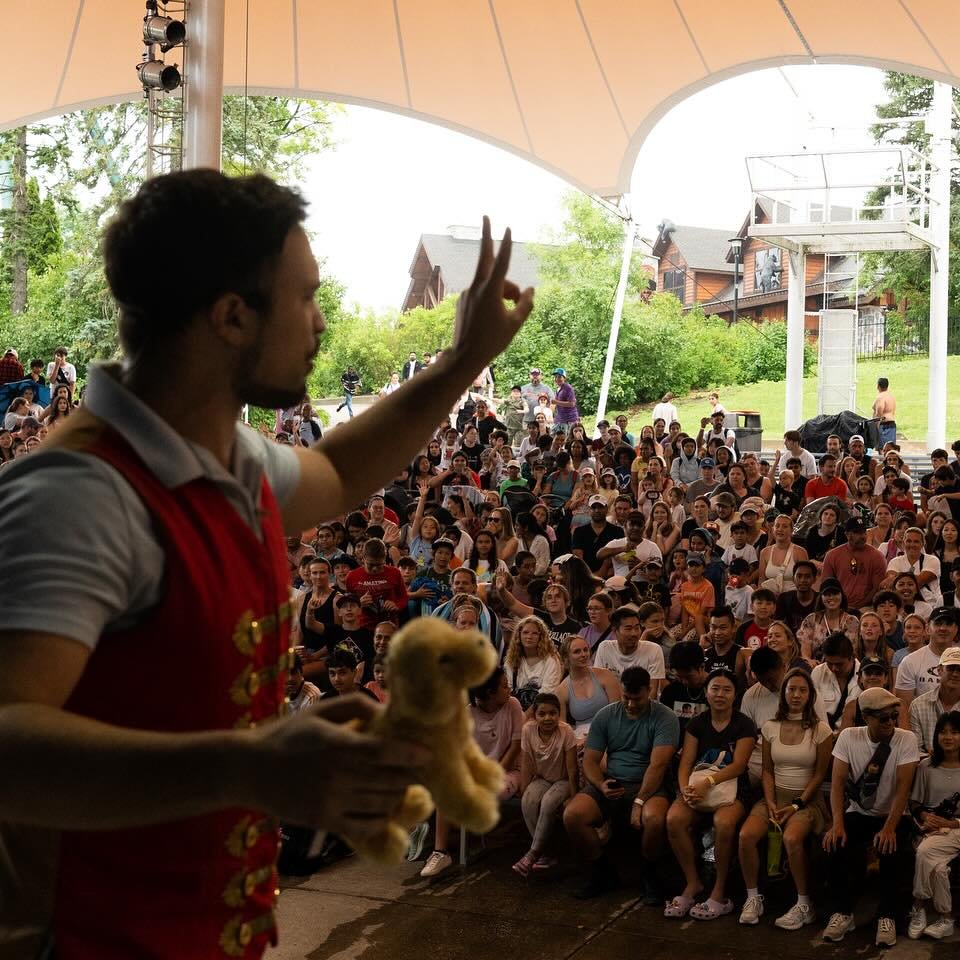 Aaron Matthews performs a captivating magic act in front of a large outdoor audience. Wearing a red vest, he gestures with enthusiasm while holding a small plush toy, engaging the excited crowd under a tented stage.